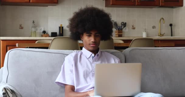Улыбающийся африканский подросток сидит на диване с ноутбуком — стоковое видео