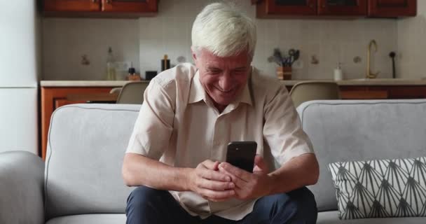 Glücklicher älterer Mann sitzt zu Hause und hält Smartphone in der Hand — Stockvideo