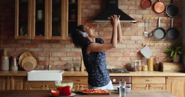 Joven mujer africana con auriculares preparando el almuerzo cantando imitando karaoke — Vídeo de stock