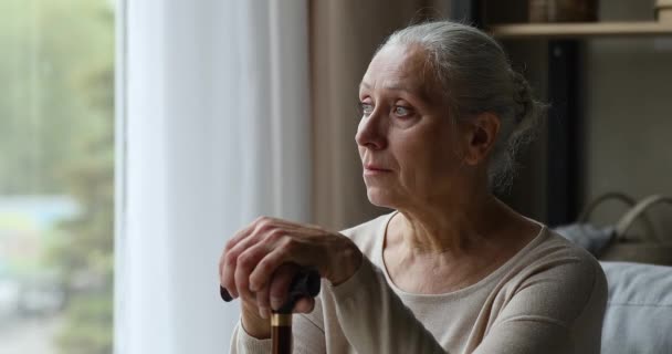 Mature woman sits on couch holds cane looks into distance — Stock Video