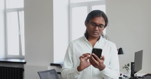 Indiase zakenman in bril met behulp van smartphone tijdens de werkdag in kantoor — Stockvideo
