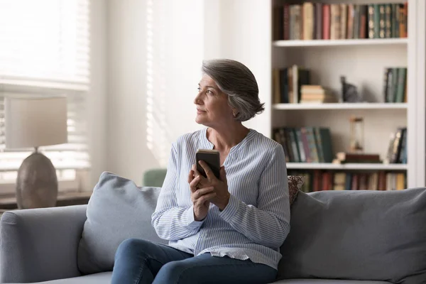 Мечтательная женщина средних лет держит в руках мобильный телефон. — стоковое фото