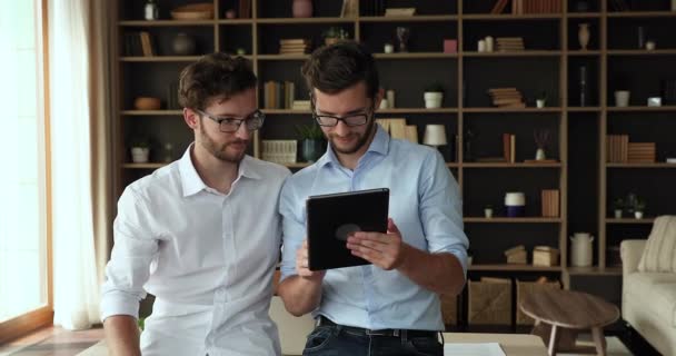 Millennial twin brothers stand by table talk looking on tablet — Stock video
