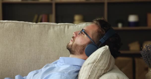 Millennial-Mann trägt Kopfhörer, die auf Couch liegen und Musik hören — Stockvideo