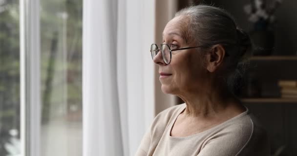 Грустная пожилая женщина вздыхает стоит у окна и смотрит наружу — стоковое видео