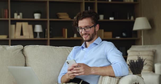 Sonriente joven relajarse en el sofá chat en el teléfono en línea — Vídeos de Stock