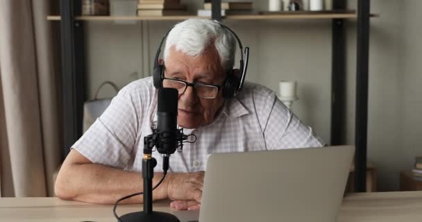 Літній чоловік сидить за столом з ноутбуком, що розмовляє мікрофоном — стокове відео
