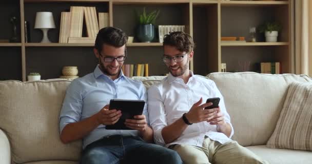 La unión de hermanos gemelos millennial relajarse en el sofá con el teléfono inteligente touchpad — Vídeo de stock