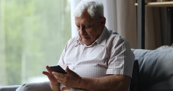 Hombre de más de 70 años se sienta en el sofá usando un teléfono inteligente moderno — Vídeos de Stock