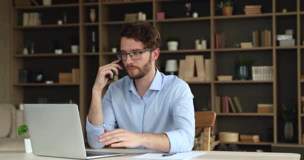 Миллениальный бизнесмен сидит за ноутбуком вести переговоры с клиентом по телефону — стоковое видео