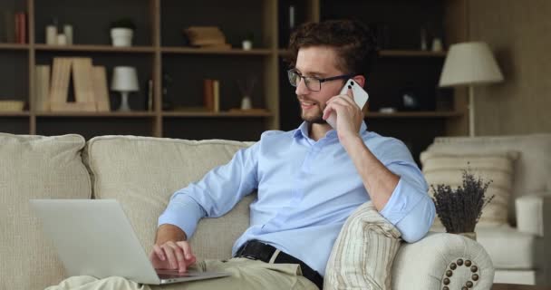 Millennial man zitten op de bank praten over de telefoon met behulp van laptop — Stockvideo