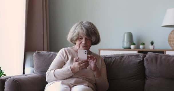 Starsza kobieta siedzieć na przytulnej kanapie za pomocą nowoczesnego smartfona — Wideo stockowe