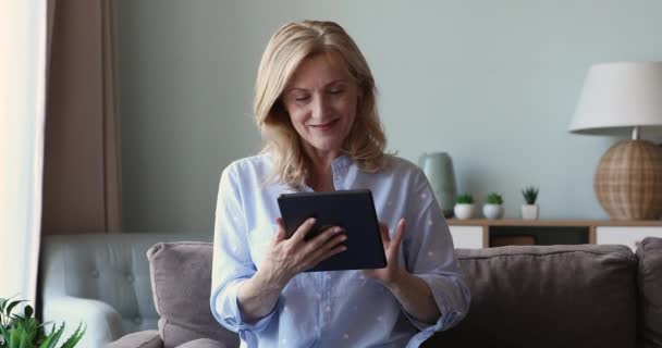 Hymyilevä houkutteleva vanhempi nainen istua sohvalla käyttää digitaalinen tabletti — kuvapankkivideo