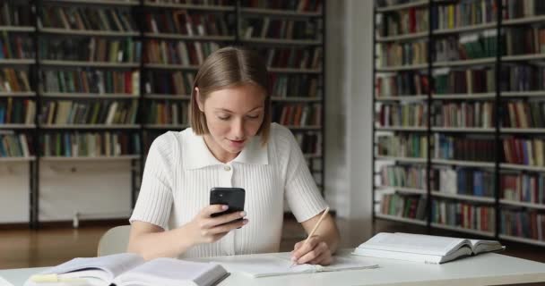 Adolescente senhora estudo na biblioteca fazer anotações a partir da tela do smartphone — Vídeo de Stock