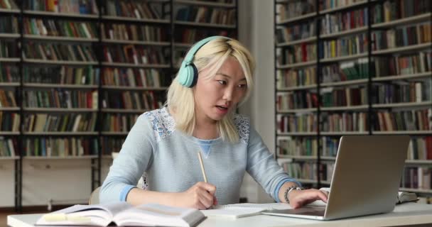 Mujer asiática usando auriculares haciendo videocall desde la biblioteca usando pc — Vídeo de stock