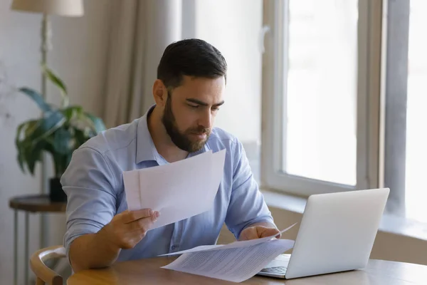 Серйозна людина сидить за столом з ноутбуком, сортуючи папери — стокове фото