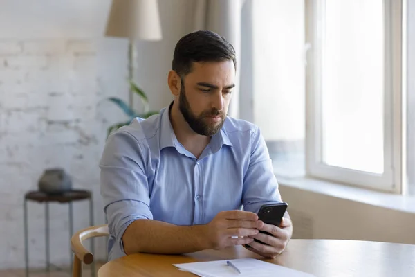 Jonge serieuze man zit aan tafel met moderne mobiele telefoon — Stockfoto