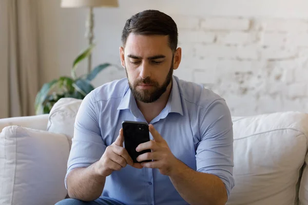 Serieuze man zitten op de bank kijken naar mobiele telefoon scherm — Stockfoto