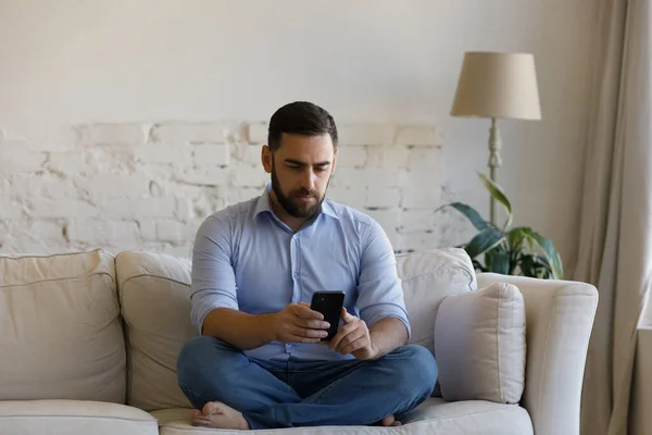 Millennial man sit on couch in living room holding smartphone — Stock Photo, Image