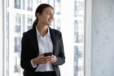 Düşüncelere dalmış mutlu bin yıllık başarılı iş kadını CEO takım elbiseli yönetici. Uzağa bakıyor. Elinde cep telefonuyla pencerenin yanında duruyor.