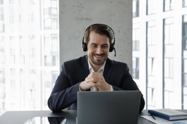 Gülümseyen Kafkasyalı genç iş adamı yönetici yönetici patron takım elbiseli mikrofon takıyor, bilgisayar ekranına bakıyor, sanal olaya katılıyor, çevrimiçi görüntülü konuşmalardan keyif alıyor..