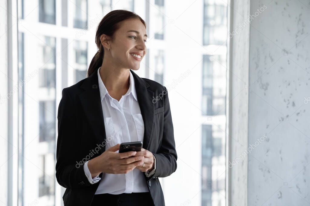 Lost in thoughts happy millennial successful businesswoman ceo executive manager in suit looking in distance, standing near window with cellphone in hands, thinking of getting pleasant news in message