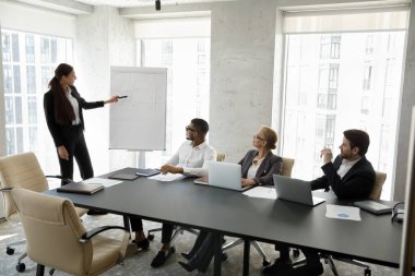 İlgili çok ırklı iş ortaklarına ya da meslektaşlarına eğitici konferanslar veren resmi kıyafetli genç bayan konuşmacı, modern yönetim odasındaki tabloların yanında duran tabloları açıklıyor.