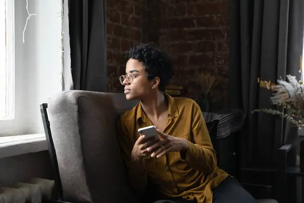 Thoughtful African Millennial Girl Glasses Holding Smartphone Looking Away Window — Stock Photo, Image