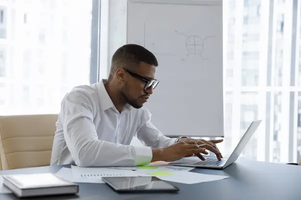 Konzentrierter Junger Afroamerikanischer Geschäftsmann Mit Brille Der Auf Den Laptop lizenzfreie Stockbilder