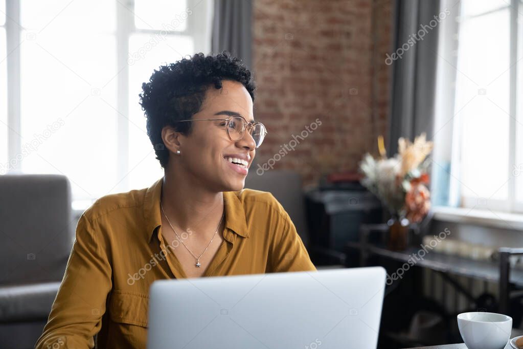 Happy positive African employee woman in glasses enjoying working at computer, looking at window away, thinking over successful project, future profit, smiling, laughing, feeling joy