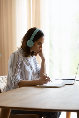 Dikey görüntü kadını kulaklık takıyor, ses dersi dinliyor, ödevi laptopla masaya oturtuyor. Kendi kendine eğitim kolay uzak öğrenimler, yeni bilgi kullanımı çevrimiçi kaynak, teknoloji konsepti