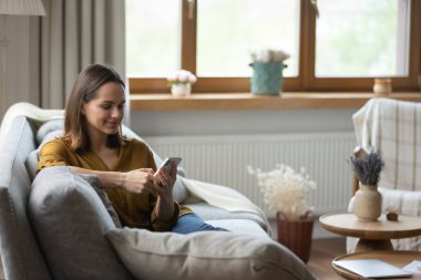 Güzel 30 'lu yaşlardaki genç bir kadın akıllı telefon gülümsemesi tutuyor. Hoş mesajlar okuyor, yeni mobil uygulamaların keyfini çıkarıyor. Kanepede tek başına dinleniyor. Malları internetten almak, sipariş vermek, evden e-alışveriş yapmak.