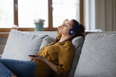 Kaygısız kadın cep telefonuyla evde uzanıyor kulaklık takıyor favori pisti kullanıyor mobil uygulama, kablosuz teknoloji, boş konsept ile dijital müzik hizmetleri kullanıyor.