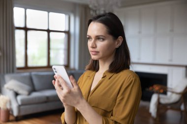 Düşünceli, düşünceli, çekici bir kadın lüks bir oturma odasında duruyor modern akıllı telefonu elinde tutuyor uzaktan bakıyor, düşünenler mesajı alıyor, cevap üzerinde düşünecekler. Teknoloji kavramı
