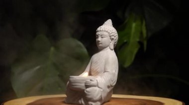 Yanan mum ateşi ve su buharı olan Buda heykeli. Tahta masa yüzeyindeki taştan yapılmış Buda doğal egzotik orman yeşil arka planda canavar bitkisi ile birlikte..