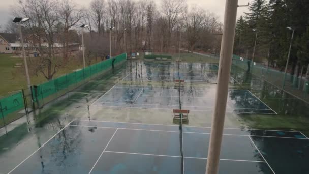 Aerial cinematic top view and movement up behind light tower of the tennis court. Slow rising up above empty tennis stadium. Rainy summer day. — ストック動画
