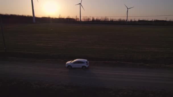 Aerial shot of car SUV driving at the golden hour by rural road. Vehicle travels near beautiful farming fields and electric wind turbines surrounded by trees. Alternative energy, agriculture concept. — Stock Video