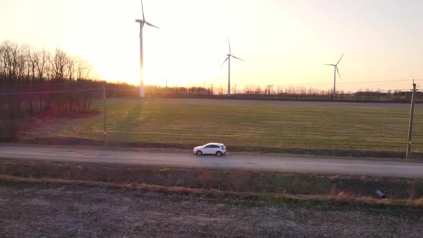White car drives on the electric wind turbines background surrounded by trees. Vehicle drives along farming fields with windmills farms at golden hour evening. Alternative energy concept. — Video