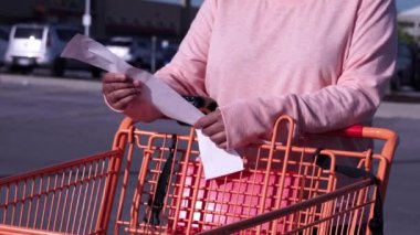 Market arabasının yanındaki kadın marketin yakınındaki kağıt fişini kontrol ediyor. Müşteri ya da alıcı yazılı faturada gıda fiyatlarını okudu. Gıda maliyetleri enflasyon krizleri ve istikrarsız ekonomi. Pahalı bir market..