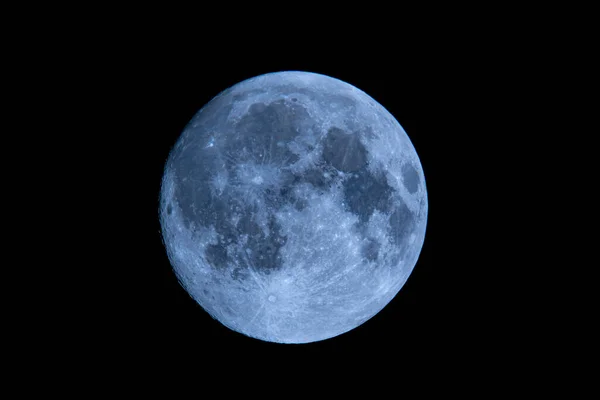 Blue full moon on dark black sky background, shot from surface of earth with very long telephoto lens. Close-up of moon glowing at clear sky.