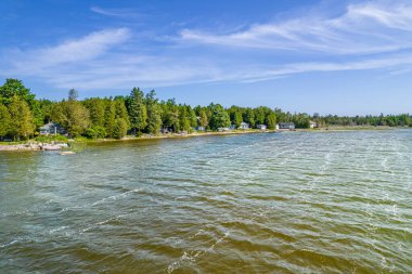 Yazın sıcacık manzarası, sahilde evler ve ağaçlar, su ve mavi gökyüzü. Balıkçı teknesinden görüntü. Kulübeler ve kiralık balıkçı kulübeleri ve özel mevsimlik eğlence.