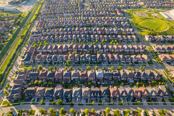 Warm summer Golden Hour evening and middle class residential houses in very geometrical setting pattern close to each other. Green grass front yards and backyards. Car roads and pedestrian walkways. 