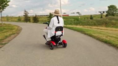 Tekerlekli sandalyede hareket edebilen scooter kullanan ve akşamları yaz parkından zevk alan bir kadın. Hareket sorunu olan tasasız bir kadın elektrikli tekerlekli sandalyede scooter kullanıyor..
