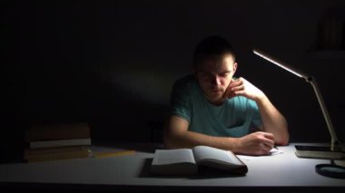 Genç bir adam ışıksız bir masada oturuyor, ve bir kitap okuyor, sınava hazırlanıyor, ve materyalleri ezberliyor, mavi bir tişört giyiyor, odada ışık yok.