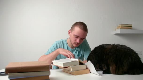 若い男がテーブルに座って本を読み 彼は試験の準備をし 材料を記憶し 彼は青いTシャツに身を包んだ 暗い猫は彼の隣に座っている — ストック動画