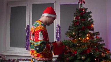 Dekorasyon ve ağaç yapan genç bir adam, Noel süveteri ve şapkası giyiyor, yanında bir yılbaşı ağacı, bir Noel ve yeni yıl kavramı var.