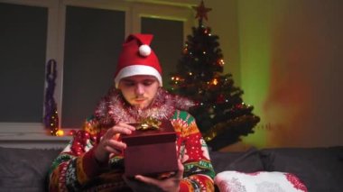 Genç bir adam kanepeye oturur ve ışık saçan bir hediye açar, Noel süveteri ve şapka giyer, yanında bir yılbaşı ağacı, bir noel ağacı ve yeni yıl konsepti vardır.