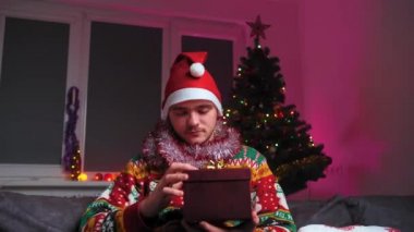 Genç bir adam kanepeye oturur ve ışık saçan bir hediye açar, Noel süveteri ve şapka giyer, yanında bir yılbaşı ağacı, bir noel ağacı ve yeni yıl konsepti vardır.