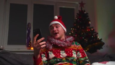 Genç bir adam kanepede oturuyor ve videoyla konuşuyor, Noel süveteri ve şapkası giyiyor, yanında bir yılbaşı ağacı, bir noel ağacı ve yeni yıl konsepti var.