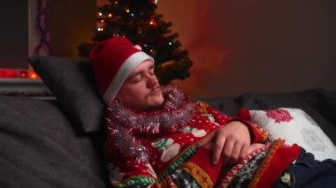 Genç bir adam kanepede uyuyor, yorgun, Noel süveteri ve şapkası giyiyor, yanında bir yılbaşı ağacı, bir Noel ve yeni yıl konsepti var.